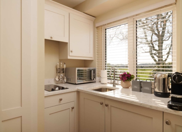 A modern kitchen with a large island, white cabinetry, stainless steel appliances, pendant lighting, and hardwood floors. A cozy seating area is visible in the background.