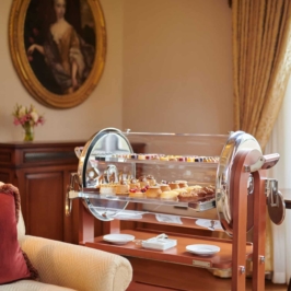 An elegant dessert trolley showcasing diverse, intricately designed cakes and pastries inside a luxurious setting, with ornate decor and rich wooden paneling in the background.