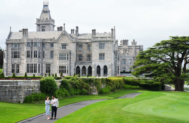 Scenic view of an elegant manor house surrounded by lush greenery, with a person cycling on a pathway amidst a picturesque landscape under a blue sky.