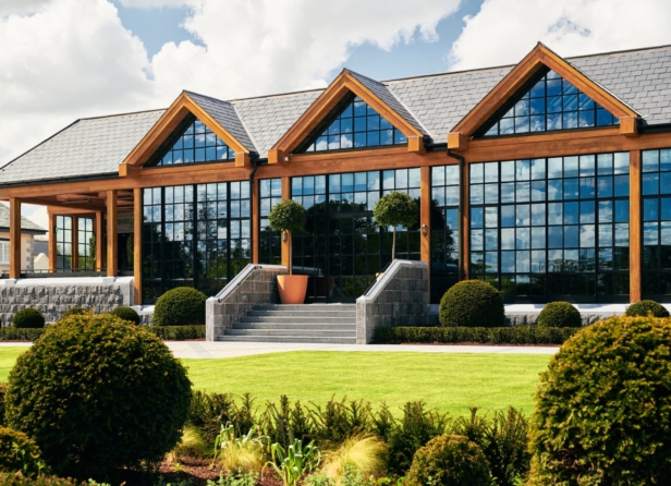 A modern wooden building with large glass windows reflecting trees, surrounded by lush greenery and a well-maintained lawn, under a partly cloudy sky.