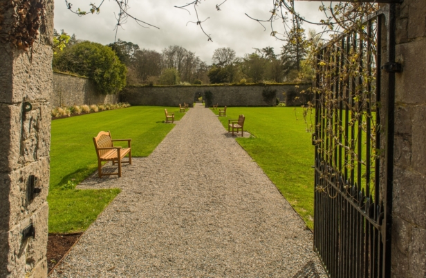 A beautifully manicured walled garden featuring a central water fountain, colorful flower beds, lush greenery, and a path leading through the vibrant landscape under a clear blue sky.